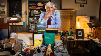 Govindjee smiles in his office, surrounded by books, instruments, plaques, and photos related to the history of photosynthesis